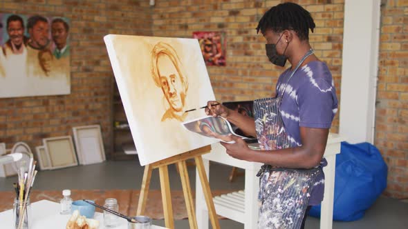 African american male painter in face mask painting from photograph on canvas in artist studio