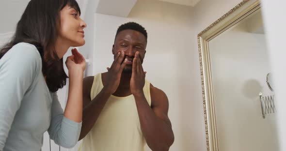 Happy diverse couple looking in mirror and using cream in bathroom