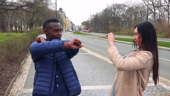 A Young Black Man Poses for a Young Asian Woman Who Takes Pictures of Him with a Smartphone