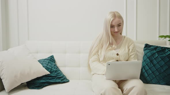 Pretty Caucasian Young Woman Working with Laptop Sitting on White Couch in Her Modern Living Room