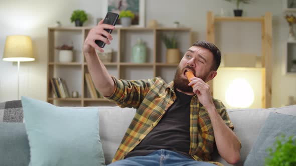 Young Man Sitting at Home on Sofa Eating Carrot and Taking Selfie