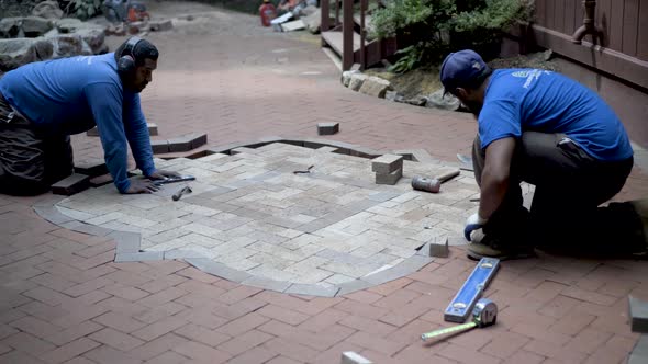 Two stone masons work on brick paver herringbone design emblem, fitting and marking bricks for cutti