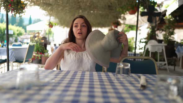 Exhausted Tired Young Woman Shaking Straw Hat As Fan Sighing Sitting in Tropic Sidewalk Cafe on