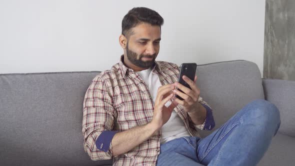 Young Attractive Indian Man Sitting on Sofa Holding Cellphone at Home