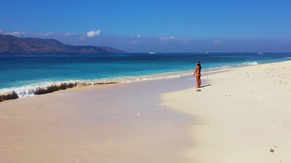 Girl in bikini and snorkel set walking on white sand of exotic beach alongside beautiful blue turquo