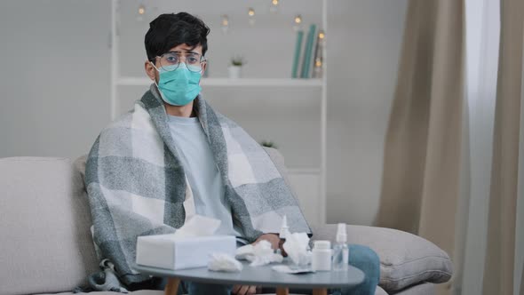 Sick Arab Man in Medical Face Protective Mask Sitting on Couch at Home Ill Guy Wrapped in Plaid