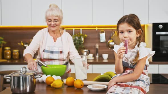 Granny Putting Flour and Egg Into Bowl Mixing It By Whisk Little Granddaughter Sitting on Table