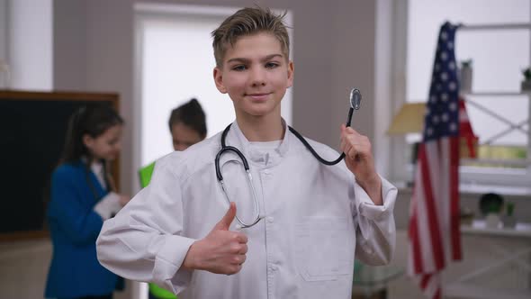 Happy Boy Showing Stethoscope Gesturing Thumb Up Looking at Camera Smiling with Blurred Friends at