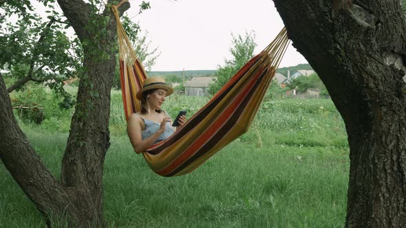 Cute woman in hat and earphones listening to music, singing the song and using mobile in hammock