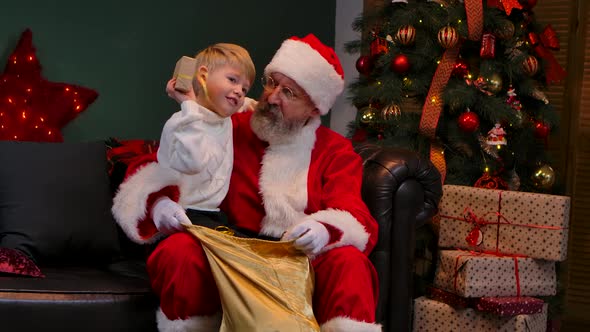 Cute Child Sits on the Lap of Santa Claus and Chooses a Gift in a Bag for Himself