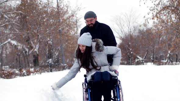 Joyful Man Rolls Wheelchair with Disabled Person Woman Through Winter Park Woman Picks Up Some Snow