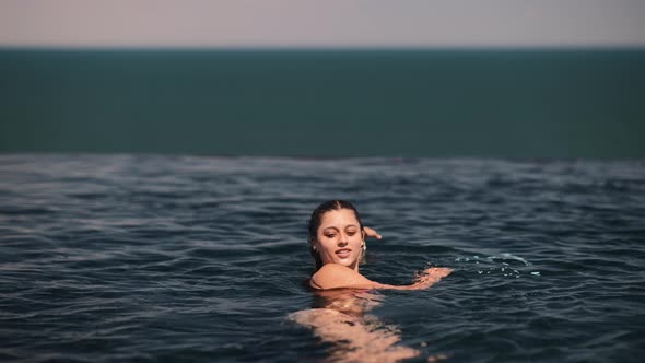 Happy Woman in Swimsuit Swimming in Infinity Pool Against Seafront