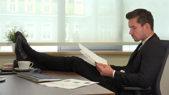 An office worker in a suit sits with legs on his desk and reads papers for work