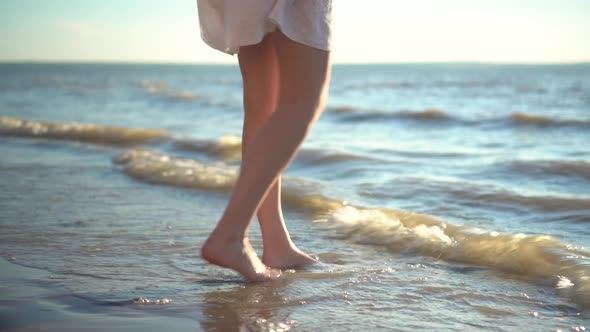 A Young Woman Stands Barefoot on the Sandy Seashore. The Girl Wets Her Feet Close-up.