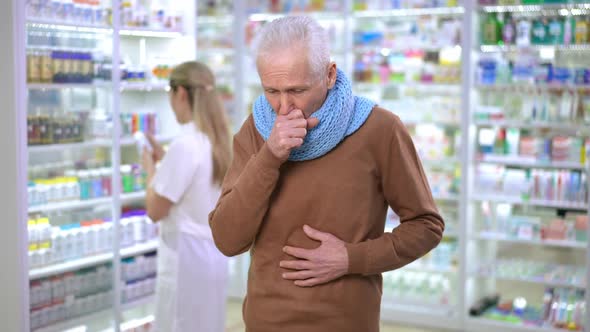 Medium Shot of Sad Ill Senior Man Coughing Standing in Pharmacy with Worried Blurred Woman Choosing