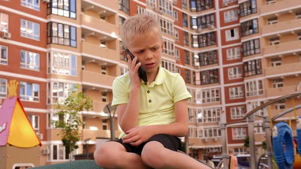 A Little Sad Boy is Talking on the Phone in the Playground