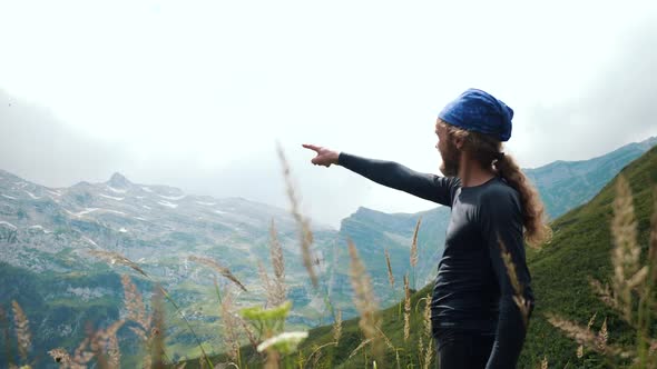 Man Hiker with Long Hair in Mountains Points with His Hand To the Mountain Peak. Mountaineer Enjoys