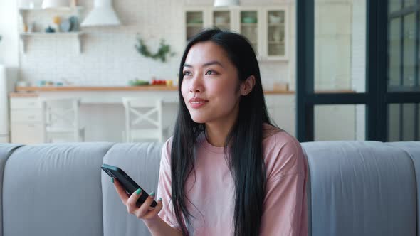 Charming and Mysterious Pensive Young Asian Woman Looking Around Thoughtfully While Sitting on Sofa