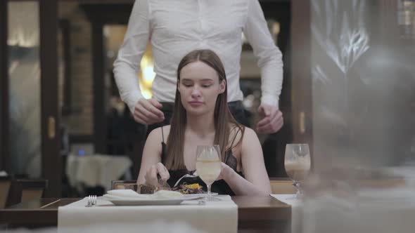 Portrait of a Beautiful Young Girl Dining in a Cafe or Restaurant. A Bearded Guy or Husband Comes Up