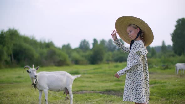 Rural Girl in Hat and Romantic Dress Posing with Goat on Countryside Field. Country Girl Walking on