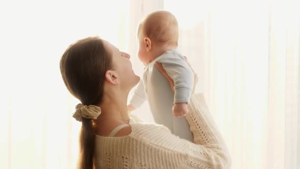 Portrait of Happy Smiling Mother Standing at Big Window and Holding Her Little Baby Son