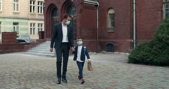 Little Child with Backpack and His Father in Medical Masks Holding Hand in Hand While Walking