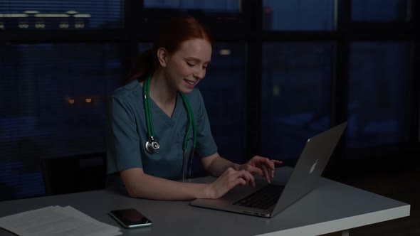 Positive Cheerful Redhead Young Female Doctor in Blue Green Medical Uniform Working Typing on Laptop
