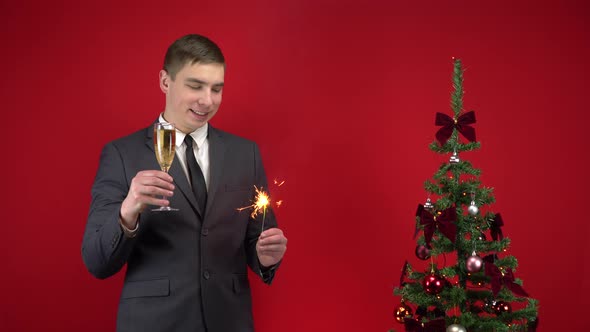 A Young Man Stands with a Glass of Champagne and a Sparkler Near the Christmas Tree on a Red