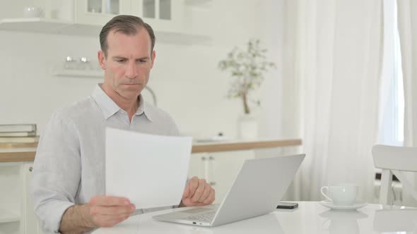 Focused Middle Aged Man Working on Laptop and Document at Home 