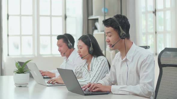 Three Asian Call Centre Agents Wearing Headsets And Typing On Computers While Speaking To Customers