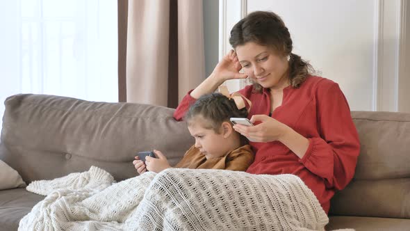 Mommy and Little Daughter Use Mobile Phones Sitting on Sofa