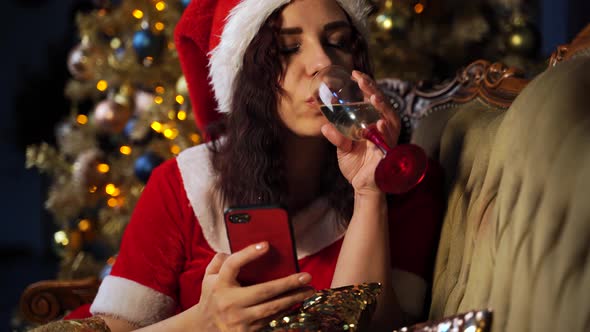 Woman in Santa Costume Browses Smartphone and Drinks Champagne Sitting on Sofa Near Christmas Tree