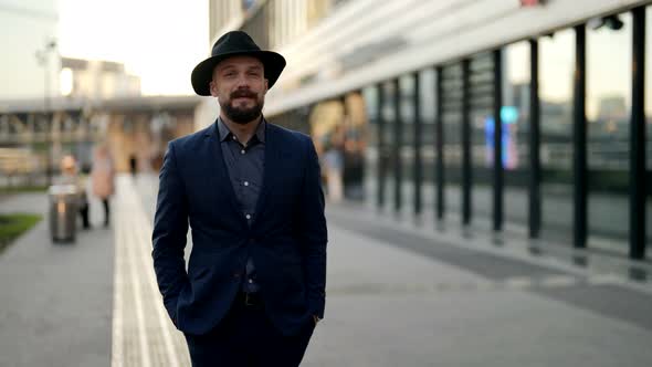 a Bearded and Moustached Man in a Hat and a Blue Jacket Walks Along a Blurry Modern Building