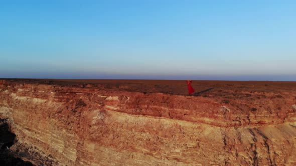 An Attractive Young Woman in a Red Dress and a Straw Hat in Windy Weather Stands on the Edge of a