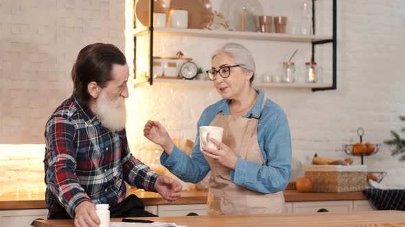 Caring Senior Woman Giving Tea To Senior Man in the Home Kitchen