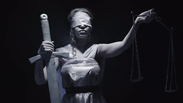 Woman Dressed and Makeup As Goddess Justice Holds Sword and Lifts Scale Standing Against Dark