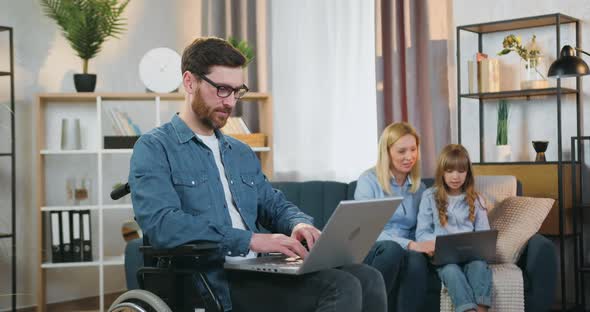 Disabled Man Working on Laptop and Looking on Camera on the Background of His Wife