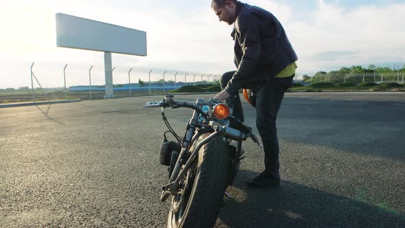 Young Stylish Motorcyclist Goes to His Custom Bobber Motorcycle on Street Road at Sunset Slow Motion