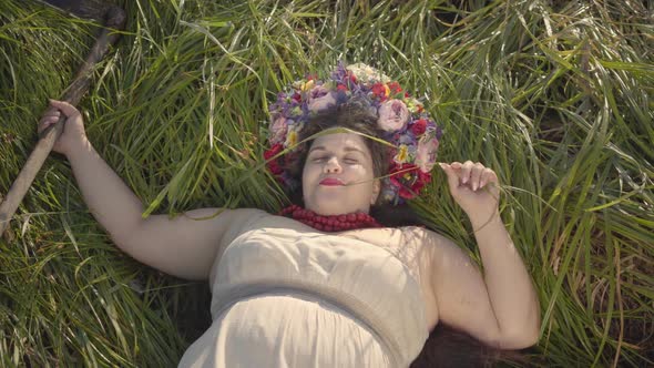 Portrait of Carefree Overweight Woman with a Wreath on Her Head Lying on the Grass in Summer Field