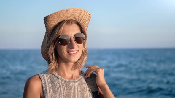 Portrait of Smiling Travel Woman in Hat and Sunglasses Posing with Backpack at Sea Sunset Background