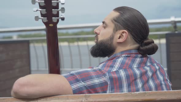 Back View Portrait of Thoughtful Young Bearded Man Sitting on Bench with Guitar and Looking Away