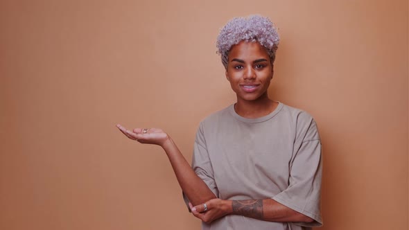 Young African American Woman Student Shows Empty Palm with Copy Space and Smiles