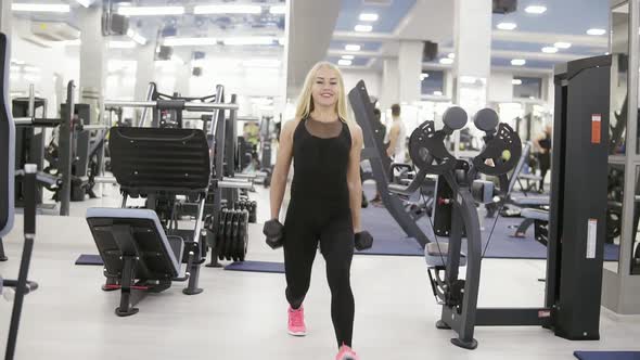 Young Attractive Female Bodybuilder Performs Squats with Dumbbells in Her Hands