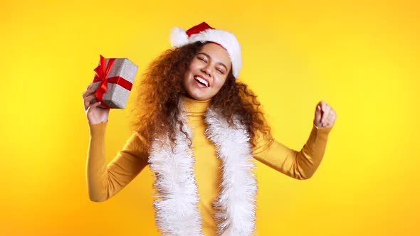 Young Pretty African Woman Dancing with Gift Box on Yellow Studio Background