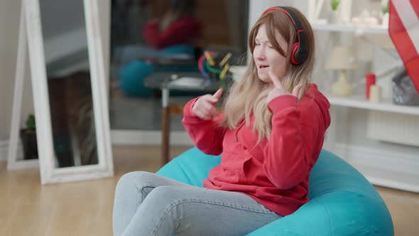 Portrait of Cheerful Relaxed Asian Teen Girl in Headphones Sitting on Bag Chair Dancing