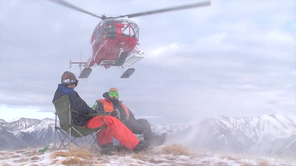 Two men sit on the top of a mountain as a helicopter drops off supplies.