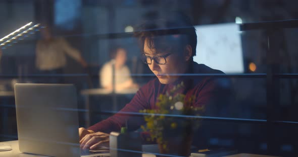 View Through Window of Young Asian Businessman Working Late at Night in Office