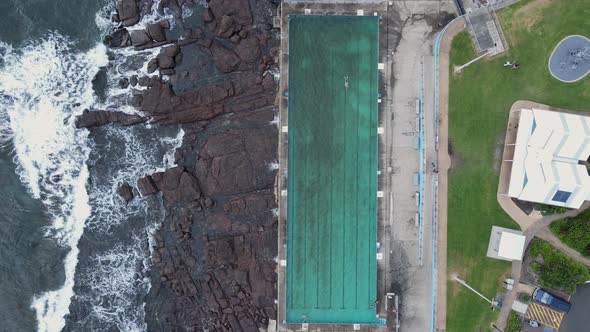 Large waves crash close to a coastal community swimming pool while people exercise. High drone view.