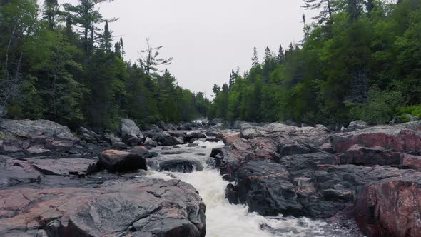 Drone footage flying low slowly over a rushing river with rapids surrounded by forest with boulders