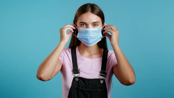 Young Woman Puts on Face Medical Mask During Coronavirus Pandemic. Portrait on Blue Background